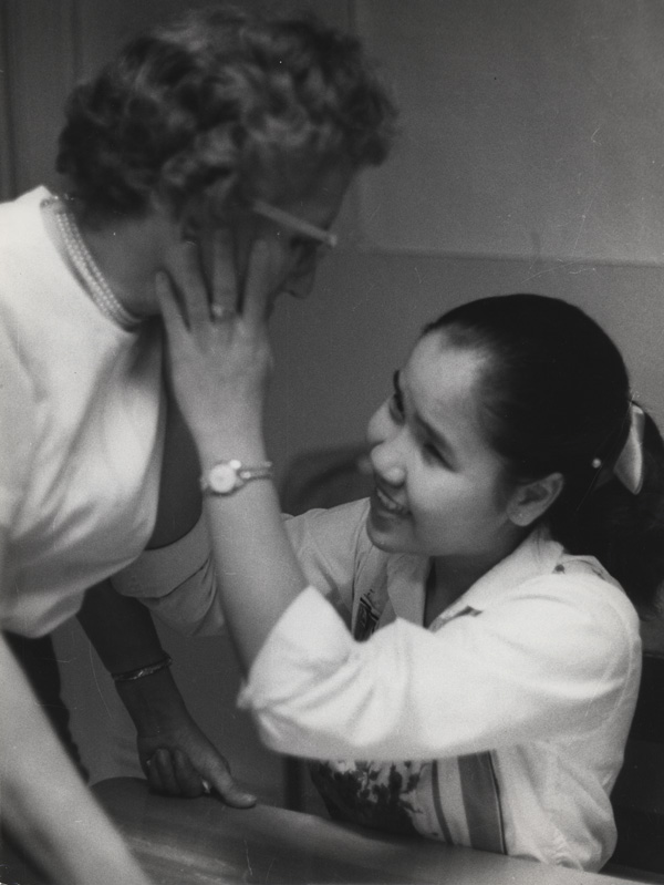 A woman stands and leans toward a seated smiling girl who is touching the woman’s right cheek.