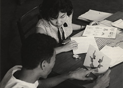 A woman observes a man seated to her right as he holds a card pointing to an image.  