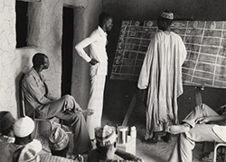 Several men are gathered in a room; one of the men is standing at a chalkboard with the instructor.