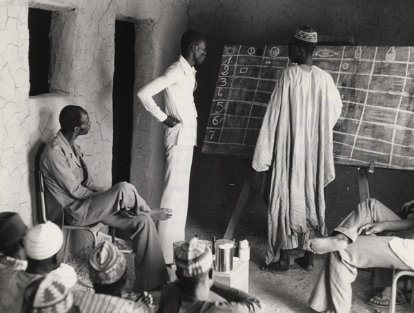 Several men are gathered in a room; one of the men is standing at a chalkboard with the instructor.