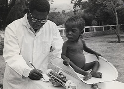 Outside, a physician weighs a child seated on a scale. 