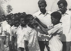 A boy puts eye drops on another boy first in line while two men look over them.