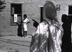 A woman stands and points to an eye chart while a person stands in the foreground taking the test. 