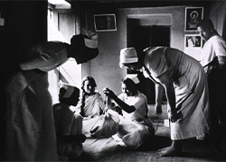 In a room, a seated woman holds a baby in a bundle hanging from a handheld scale while several other women surround and observe her.