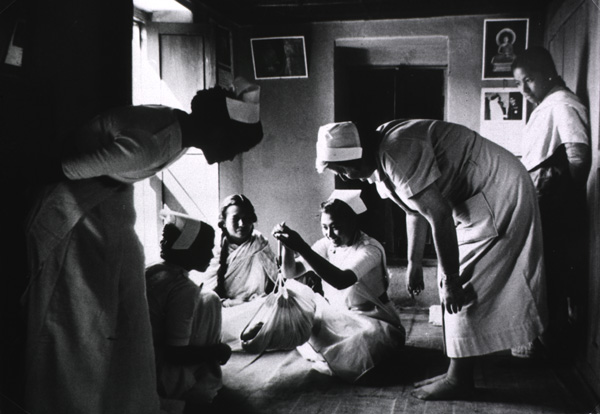 In a room, a seated woman holds a baby in a bundle hanging from a handheld scale while several other women surround and observe her.