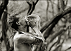 Black and white photograph of man with palm fronds around his neck blows into a conch shell during the Makahiki.