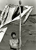 : Black and white photograph of a shirtless man underneath a t-shaped pole made of wood with fabric draped across.