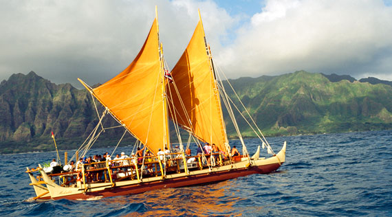 A canoe with 2 yellow sails travels across a dark blue ocean. A mountain and sky appear in the background.