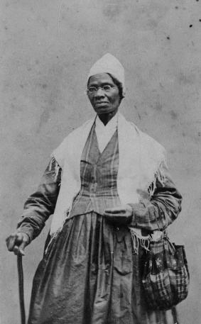 An elderly woman stands with a cane in her right hand and a bag on her left, wearing a cap and a shawl.