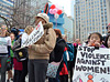 A diverse crowd of people, mostly women, stand holding signs in a plaza.