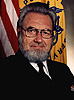 Bearded White man wearing a black military style uniform seated and looking at viewer.