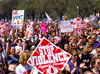 A large crowd stands on the Mall facing right, many hold signs.