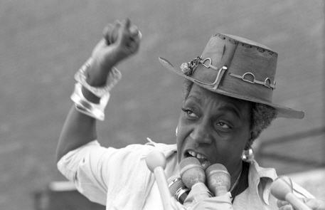 Activist Flo Kennedy with raised right arm, speaks into a bank of microphones to a crowd out of view