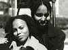 Two African American women seated and facing viewer, woman on left front leans into woman behind.