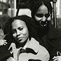 Two African American women seated and facing viewer, woman on left front leans into woman behind.