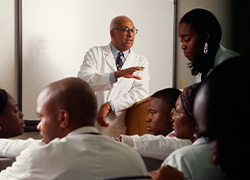 A man leans against a lectern speaking to people seated in a classroom.