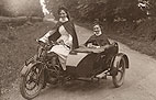 Two White female nurses in a motorcycle with sidecar. Both wear white uniforms under their cloaks.