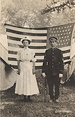 A White female nurse and a White male soldier in uniform stand in front of an American flag.