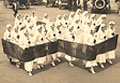 A group of White female Red Cross nurses in white march in a cross formation.