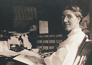 A woman at seated at a desk, turning towards her left to look at viewer.