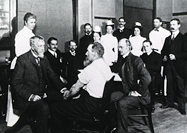 Dr. Mitchell examining a male patient in left foreground while a group of men and women look on.