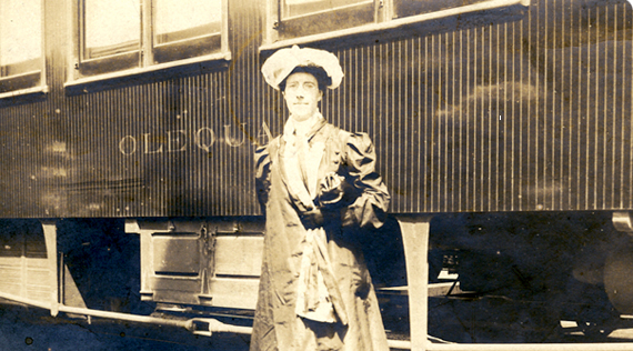 A woman in dress and hat standing in front of a train car.