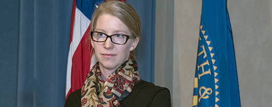 Woman wearing glasses facing forward between the US flag and DHHS flag.