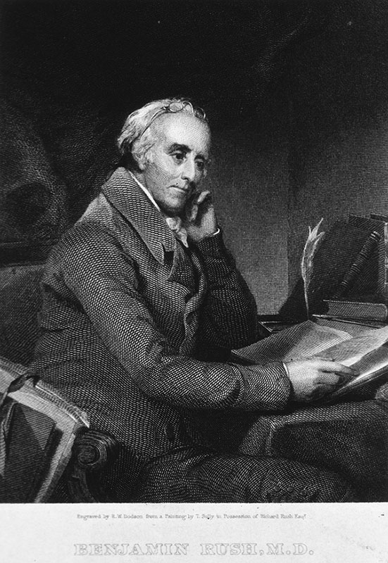 Formal portrait of a white man seated at a desk with an open book; glasses on forehead; thoughtful pose.